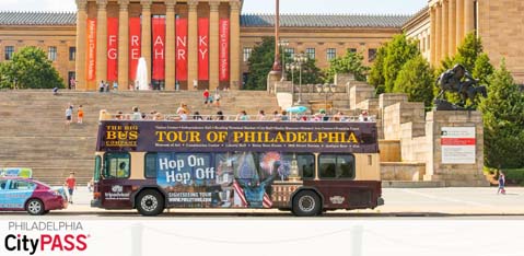 An open-top, double-decker bus branded for the Tour of Philadelphia with images of famous landmarks and the phrase  Hop On Hop Off  is in the foreground. In the background, the Philadelphia Museum of Art with its iconic steps, and columns with red banners. A statue on horseback is visible to the right, and the words  Philadelphia CityPASS  adorn the bottom of the image.