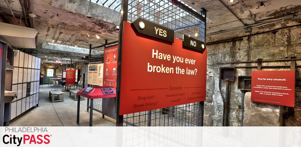 Image of an interactive exhibit at the Philadelphia CityPASS attraction. Large red and black signs ask, 'Have you ever broken the law?' with 'YES' and 'NO' options. Bullet points beneath include 'Drug Use?' and 'Drunk Driving?' Additional text touches on the rarity of never breaking the law. The setup is in an industrial-looking space with exposed ceiling and walls.