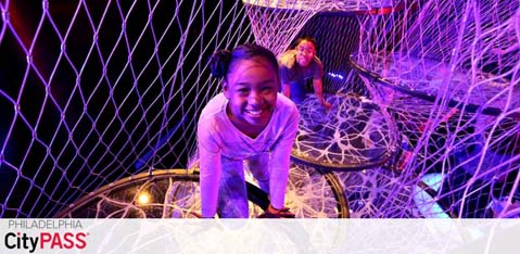 This image features two joyful children playing inside a vibrant, web-like structure. The foreground shows a girl smiling at the camera while crouched down, with colorful lighting enhancing the playful atmosphere. Behind her, another child can be seen enjoying the space. The words 'Philadelphia CityPASS' suggest this location is part of a city attraction package.