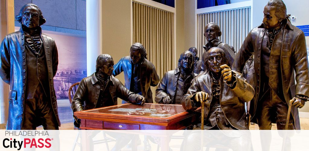 Image shows a group of life-sized, bronze statues depicting historical figures in a meeting setting. They are grouped around a table, some standing, some seated, all appearing engaged in discussion. The backdrop suggests an indoor setting with paneled walls and a faint cityscape. A logo for Philadelphia CityPASS is in the upper left corner, indicating a possible tourist attraction.