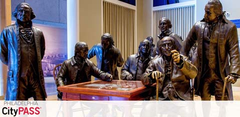 Image displays a collection of bronze statues representing historical figures gathered around a table, as if engaged in a discussion. Situated indoors, a banner above reads 'Philadelphia CityPASS'. The setting suggests a museum or educational venue showcasing American history.