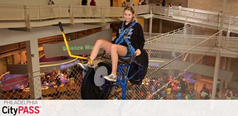 A smiling individual enjoys a bicycle ride on a high-wire attraction at the Franklin Institute Science Museum. Below, visitors are visible in the multilevel atrium. The space is bright with natural lighting. Promotional text for Philadelphia CityPASS is displayed in the corner.