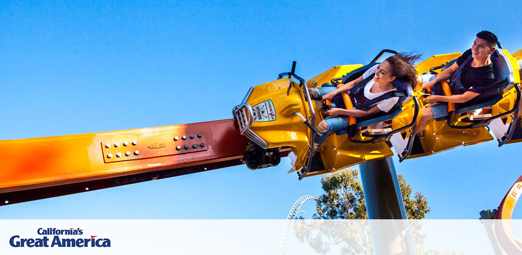 This image features a thrilling ride at California's Great America theme park. A clear blue sky serves as the backdrop for the photograph. To the right, two riders are seated in a bright yellow two-person roller coaster car that is suspended from an orange track above them. Both riders appear to be joyfully engaged in the experience, with the person on the left raising their arms high in apparent excitement, while the person on the right is holding onto the safety restraint, smiling broadly. In the background, faint outlines of trees and part of a roller coaster structure can be seen. The image conveys a sense of fun, excitement, and the adventurous spirit of the amusement park.

At GreatWorkPerks.com, we're committed to offering our customers not just excitement but also amazing savings—be sure to check out our selection for the lowest prices on tickets to experience the fun at California's Great America.