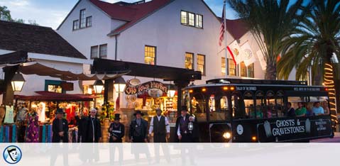 This image displays a bustling evening scene at a charming, historic location. On the right-hand side of the image, a black trolley bus with the words "Ghosts & Gravestones" written on its side is stationary on the street, suggesting it provides themed tours. The trolley embodies an older, classic design aesthetic which complements the historic vibe of the surroundings. On the left, we see a row of quaint shops with people mingling and walking on the sidewalk in front of them. The shops' architecture has a traditional style with white walls and a red-tiled roof, emblematic of Spanish colonial influence. Several flags are visible atop the buildings, waving in the dusk light. The sky is painted with soft hues of blue and purple as evening sets in, creating a welcoming atmosphere for visitors and night-time shoppers alike. At GreatWorkPerks.com, we're dedicated to ensuring our customers enjoy incredible savings on tickets to many such unforgettable experiences, ensuring the lowest prices for your next adventure.