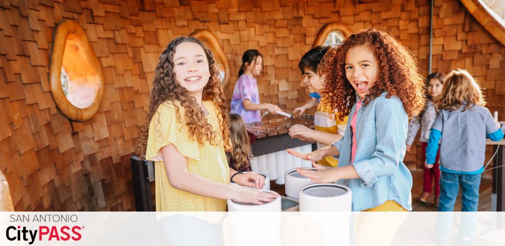 Two joyful children engage in an interactive exhibit with others in the background. The girls, one in a yellow dress and one in a denim jacket, are smiling brightly at the camera. The setting features a warm wooden interior and a CityPASS logo, suggesting a fun, educational environment.