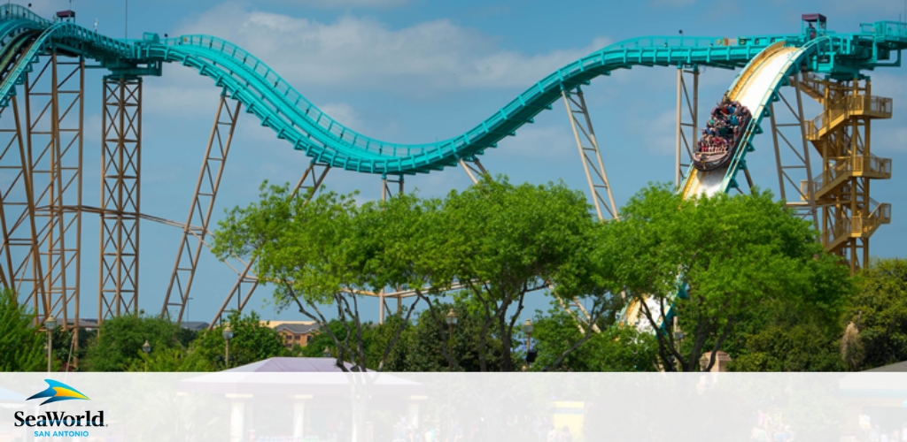 Image shows a roller coaster ride at SeaWorld San Antonio. In the foreground, there's lush greenery with a clear sky overhead. A cart filled with passengers is descending a steep track at high speed, providing an exhilarating experience. The SeaWorld logo is visible in the bottom left.