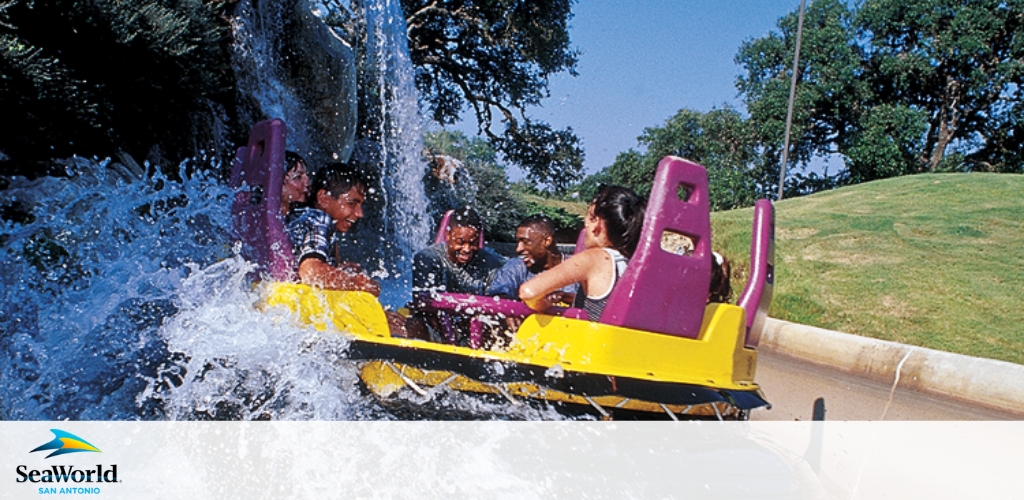 Image shows a group of excited visitors on a yellow water ride at SeaWorld San Antonio. They are splashing down a chute, surrounded by spray and water, conveying the thrill and fun of the attraction. The SeaWorld logo is visible in the corner.