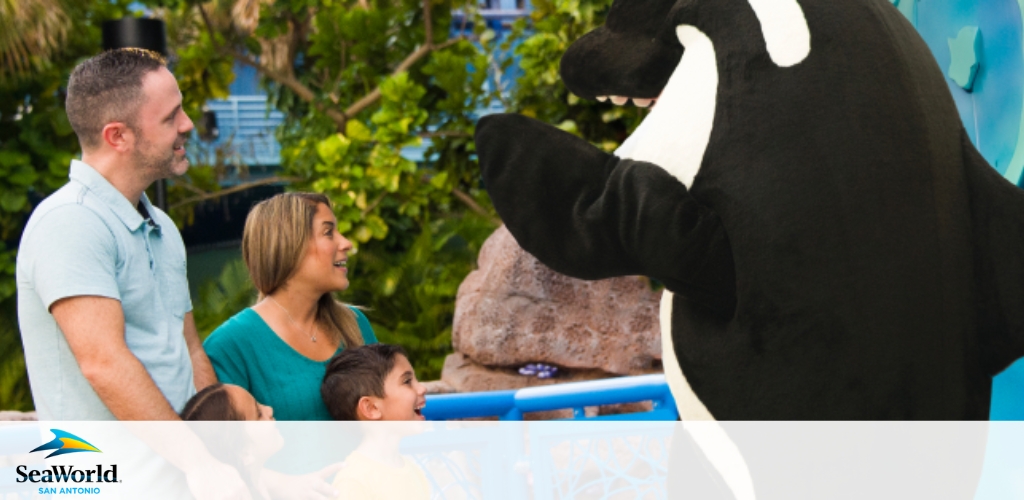 Image shows a happy family interacting with a costumed character resembling an orca at SeaWorld San Antonio. A man, a woman, and a child are smiling and engaged with the friendly character who is positioned to their right. Natural greenery and park branding are visible in the background.