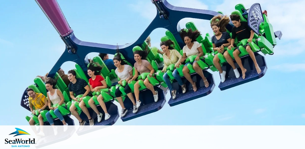 This image shows a group of excited riders on a green and blue amusement park swing ride mid-air, with lively expressions and their feet dangling. The SeaWorld San Antonio logo is visible in the corner.