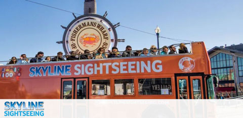 This image showcases a bright day under a clear blue sky where an open-top, double-decker Skyline Sightseeing tour bus is filled with passengers. The bus is partially framed by an iconic sign indicating the location to be Fisherman's Wharf, which is a popular tourist attraction. The passengers appear joyful and engaged as they take in the sights. In the background, there's a glimpse of a building with large windows and a few other structures indicative of an urban environment. With GreatWorkPerks.com, enjoy spectacular experiences at the lowest prices – don't miss out on great discounts on tickets to your next adventure!