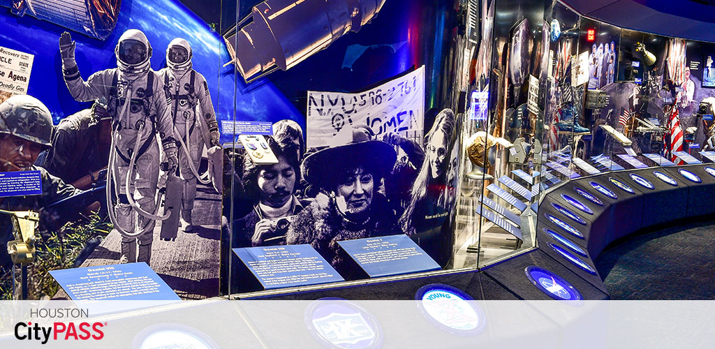Exhibit space featuring large photos of astronauts, historical moments, and interactive displays. Text panels provide information, and a curved wall of images highlights significant events. The Houston CityPASS logo is visible.