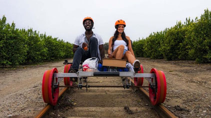 Description: This image shows two individuals riding a pedal-powered rail vehicle on a set of train tracks. The person in the front is wearing a black shirt and jeans paired with a dark colored helmet, sporting a joyful smile. The person in the back, dressed in a light-colored tank top and shorts, is similarly donned in a bright helmet and also shares a cheerful demeanor. Both riders enjoy their outdoor adventure surrounded by lush greenery on both sides of the track, indicating a rural or park setting. The rail vehicle features prominent red wheels, a simple metal frame, and a small platform for resting their feet or placing items. It seems to be a cloudy day, giving the scene a soft, natural light without harsh shadows.

At GreatWorkPerks.com, we're dedicated to bringing you the joy of activities like this rail riding adventure at the lowest prices, ensuring you get your tickets with great savings!