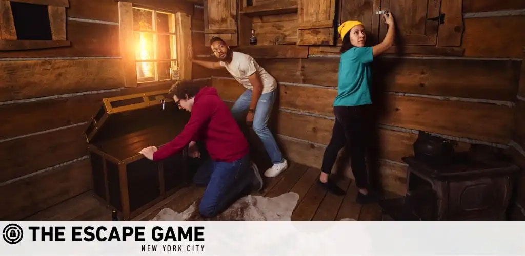 In the photo, three people are engaged in an escape room challenge. They are in a room with rustic wooden walls, working together to find clues. One person kneels to investigate a chest, another crouches with focus, and the third stands reaching for something high on the wall. The ambiance is adventurous, suggesting teamwork and discovery as they search for their next lead. The logo reads 'The Escape Game New York City'.