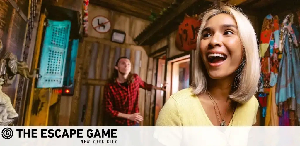 Image shows two people enjoying an escape room experience at The Escape Game in New York City. On the right, a joyful woman with short blond hair in a yellow top smiles widely. On the left, a person in a red plaid shirt examines the room, filled with various props and a rustic vibe. The location  New York City  is referenced.