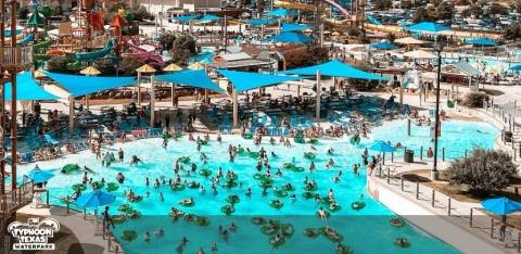 The image shows a bustling water park on a bright day. A large wave pool is at the forefront, with people enjoying the water amidst gentle waves. Blue umbrellas and loungers are scattered along the poolside. The park's vibrant atmosphere is highlighted by various slides and attractions in the background, with landscaping and walkways throughout.