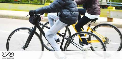 Description: This image showcases two individuals engaged in outdoor cycling. The person in the foreground is riding a black bicycle, captured from the waist down. They are wearing dark blue jeans and a puffy black coat, which suggests a cool climate. Meanwhile, the second person, partially visible and shown from the waist down, rides a yellow and black bicycle and is clad in white pants with a black stripe and a black jacket. Both riders are wearing sneakers, and their movements have been captured in such a way that conveys motion, likely indicating they are moving at a brisk pace. The background is blurred with hints of greenery and a roadway, suggesting a park-like or urban setting for this activity. The lower left corner of the image contains a logo with the initials "U B" followed by the words "UNLIMITED BIKING." 

At GreatWorkPerks.com, we're dedicated to bringing you the best outdoor experiences with the lowest prices guaranteed. Don't miss out on incredible savings - find your next adventure and grab your tickets today!