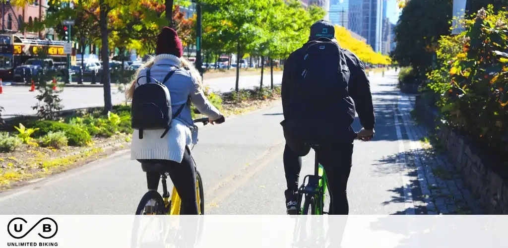 This image features two individuals engaged in a leisurely bike ride along a paved path on a sunny day. The person on the left is riding a yellow bike and is dressed in a light jacket, jeans, and a burgundy beanie, accessorized with a dark backpack. The person on the right rides a bike with vivid green accents and is wearing dark clothing and a black backpack with visible white lettering. They both appear to be pedaling at a calm pace. In the background, we can see the changing colors of trees foliage, indicating autumn season, as well as urban elements like a busy street with a city bus and traffic cones. The bottom left corner of the image contains the logo "U B Unlimited Biking," suggesting a biking service or promotion. At GreatWorkPerks.com, we’re committed to offering the lowest prices available, ensuring our customers enjoy significant savings on tickets and experiences.