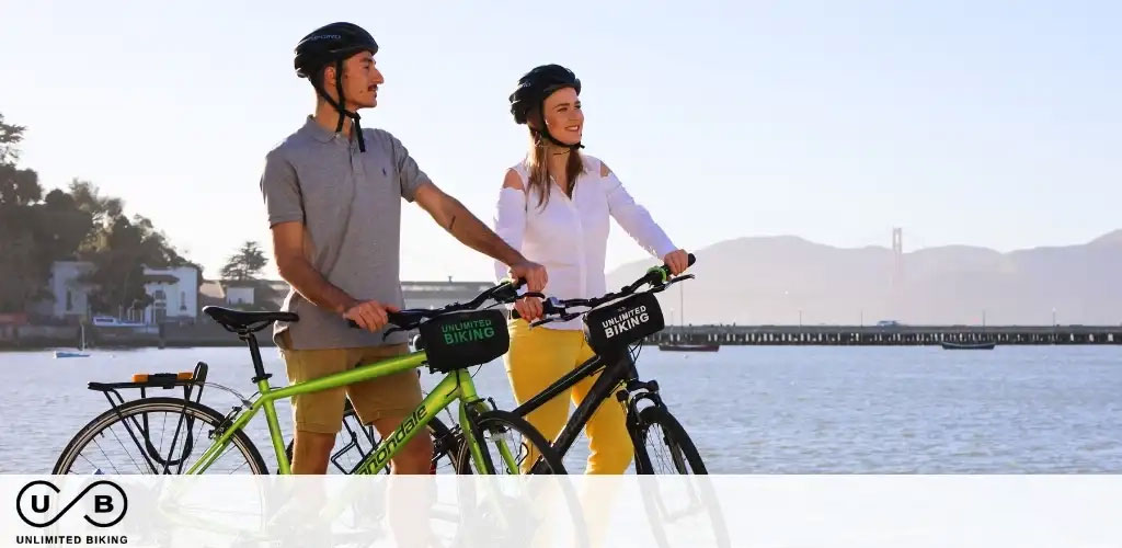 Image Description: This image presents a cheerful man and woman standing side-by-side with bicycles against a scenic backdrop featuring a calm water body and a clear sky. The man is on the left, wearing a helmet, a grey polo shirt, and dark pants. He is holding onto a green bike with the text "UNLIMITED BIKING" displayed on the frame. The woman is on the right, also sporting a helmet and a white long-sleeved shirt with light pants, holding a bike identical in branding but yellow in color. Both individuals are looking towards the horizon with content smiles. In the distance, a bridge spans across the water and gentle hills under a bright sky complete the tranquil scene.

At GreatWorkPerks.com, we’re committed to offering you the same joy and serenity as biking by the water with the added bonus of exceptional savings. Shop with us for the lowest prices on tickets and discover exclusive discounts!