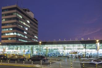Entrada do Aeroporto Internacional Jorge Chavez ao anoitecer com carros passando em frente deixando rastro de luz