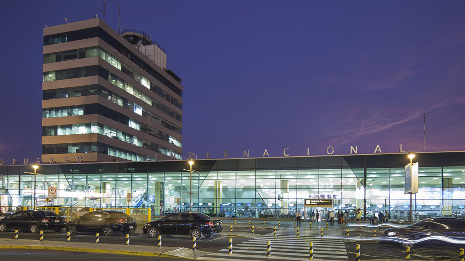 Entrada do Aeroporto Internacional Jorge Chavez ao anoitecer com carros passando em frente deixando rastro de luz
