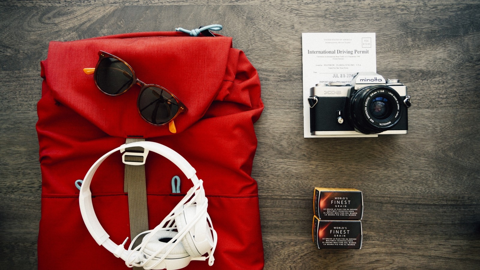 Foto aérea de uma mochila, óculos, fone de ouvido, camera, documentos e filme sob uma mesa. Photo via Visual hunt