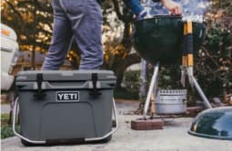 A YETI cooler sits in the foreground while a person uses a smoking kettle grill in the background.