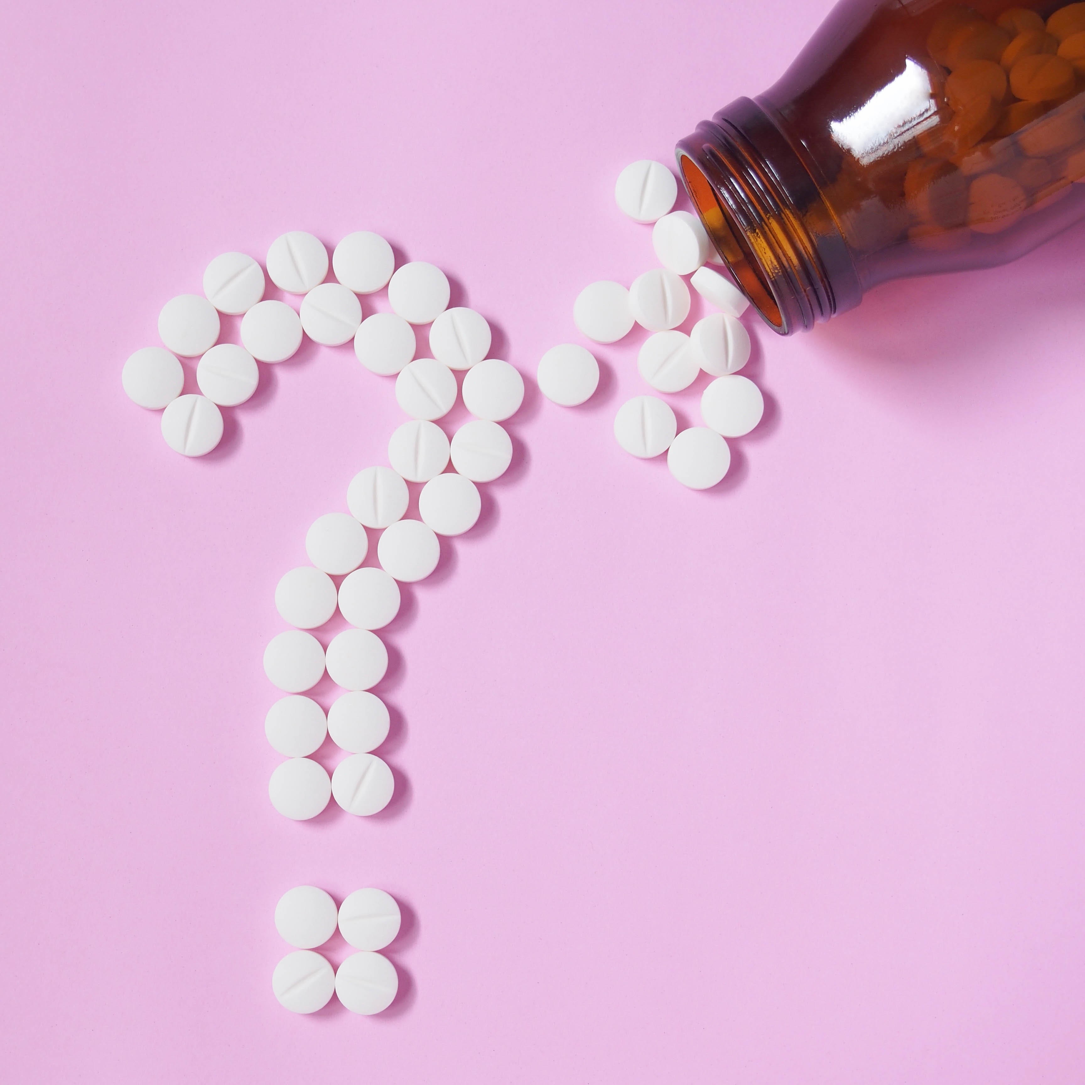 A pill bottle spills small, round tablets in the form of a question mark