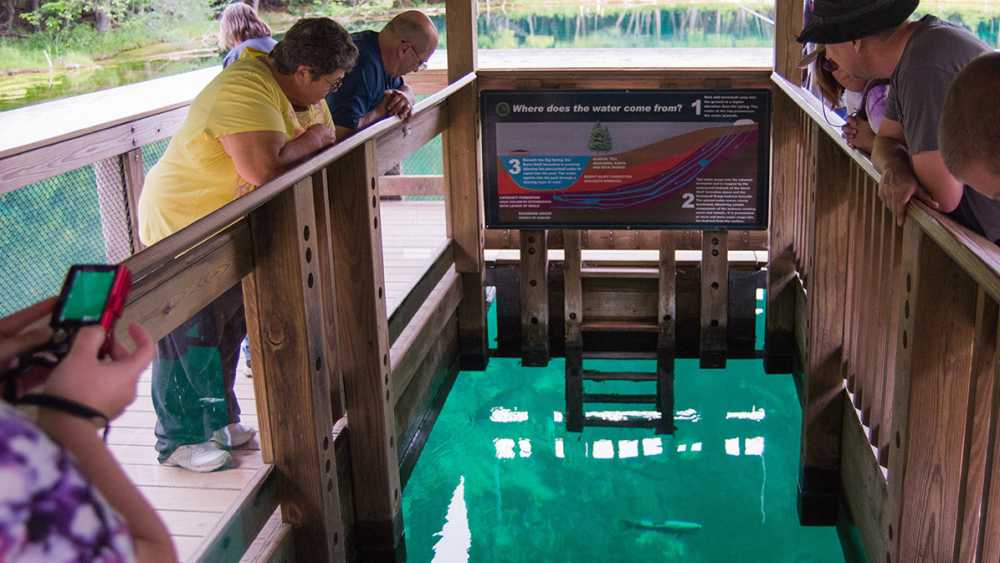 People look over the interior raft railing to view fish swimming in Kitch-iti-kipi spring. People look over the interior raft railing to view fish swimming in Kitch-iti-kipi spring.
