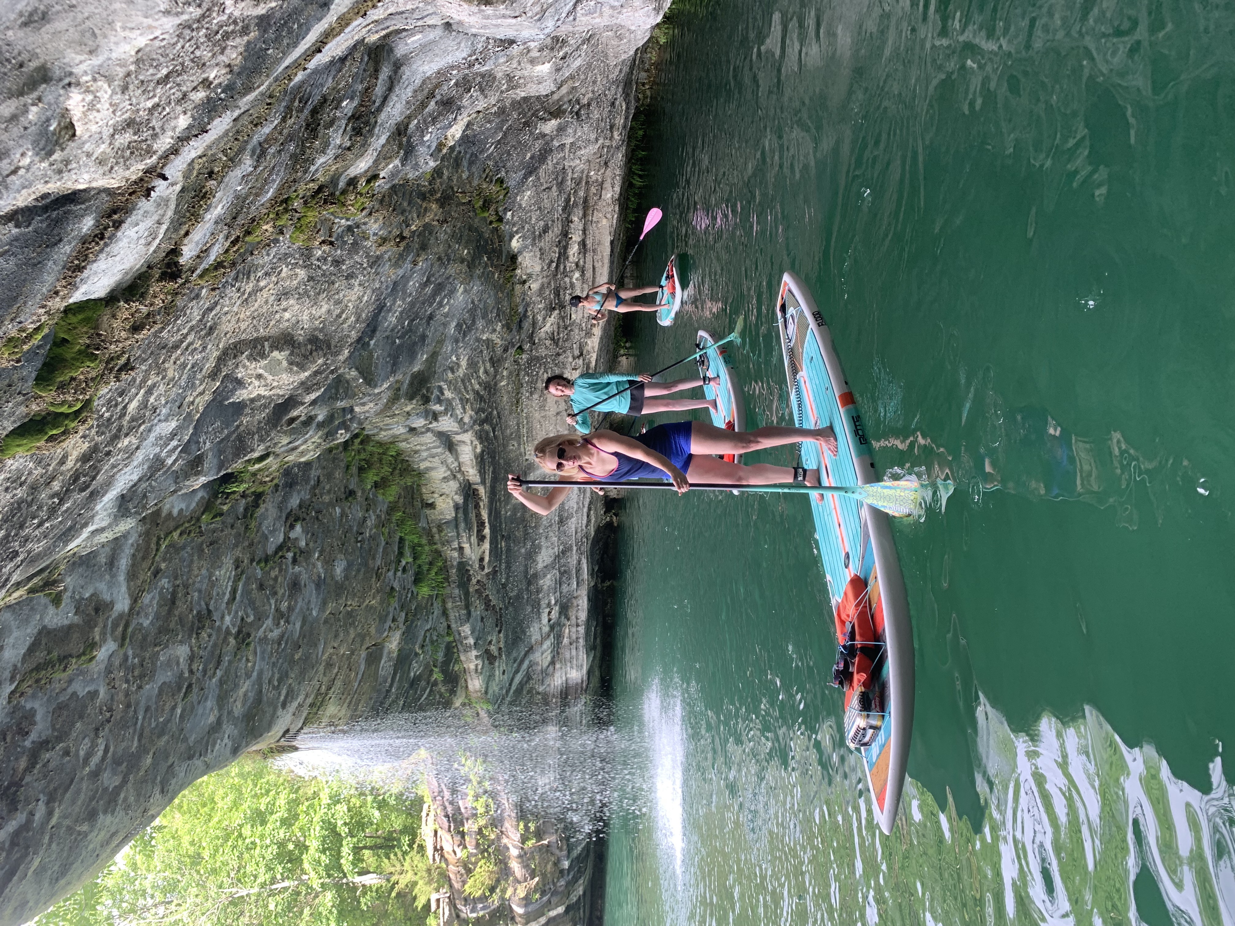 Three women standing up on light blue paddleboards. Water is below them. A limestone bluff on their left with a small waterfall in the background. Paddle Boarding to Waterfalls 