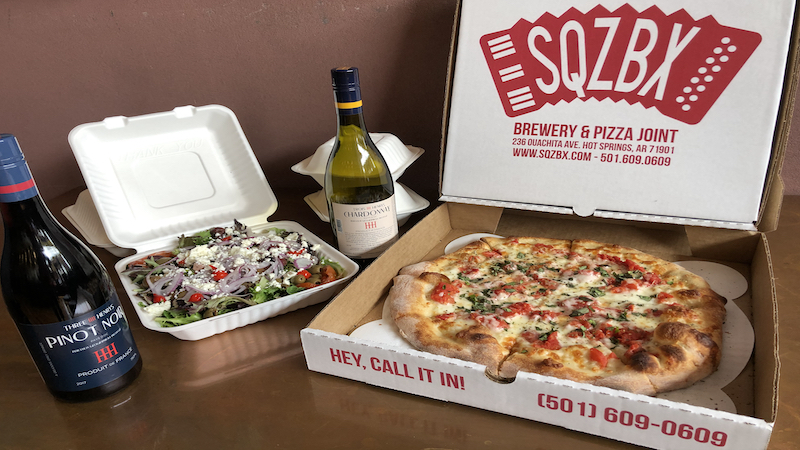 A to-go box of SQZBX pizza, salad and two bottles of wine sit on a table nicely arranged. 