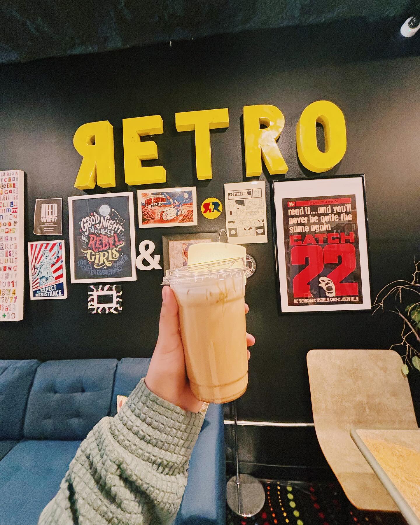 Hand holding an iced coffee in front of a black wall with artwork.