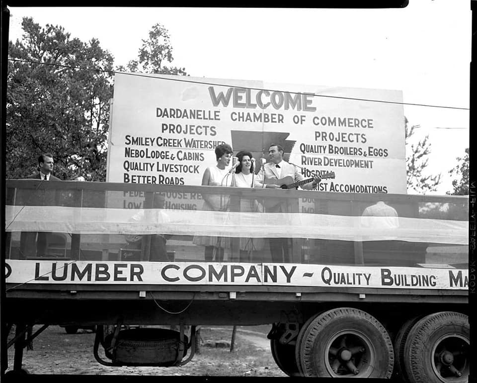 Black and white photo of trailer stage with three musicians in front of a large sign. 