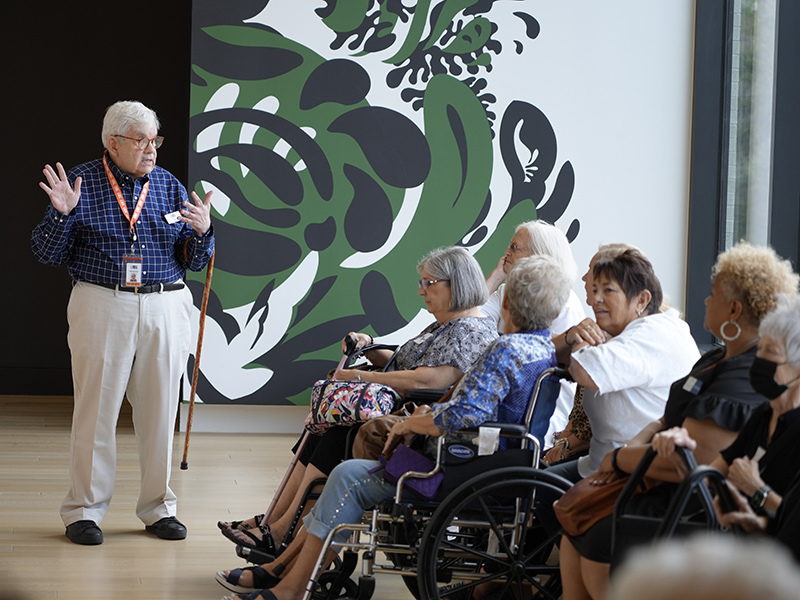 A museum guide speaking to a group of elderly visitors, some in wheelchairs, in front of a large abstract black and green artwork.
