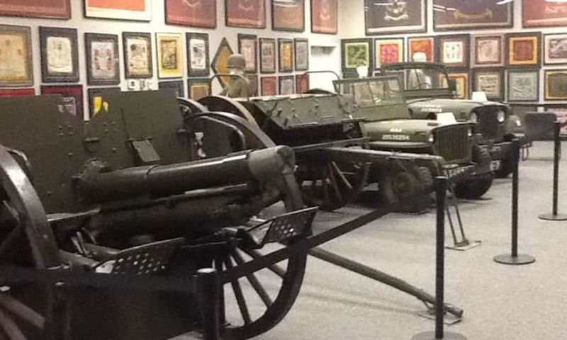 Two, original military jeeps with white numbers are on the right side of the image. Another piece of military equipment with wheels is on the left side. Framed artifacts hang on the walls.