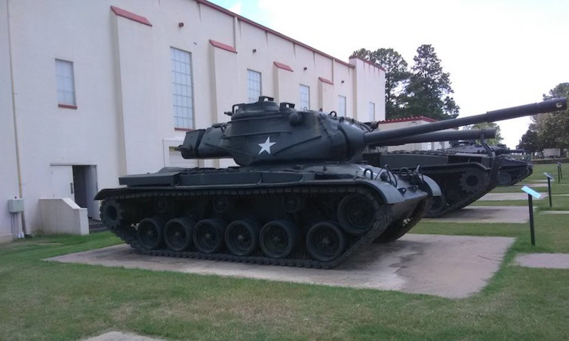 Military tanks are on display outside of a white building. A M-47 Patton tank with a white star is at the front of the image.