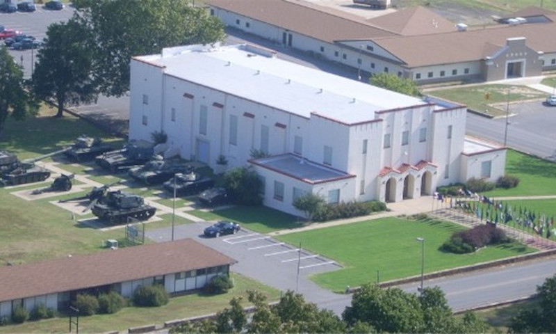 Aerial of of white building. Green grass surrounds the museum while several flags line the walkway leading to the entry. Ten military tanks are on the left of the museum. 