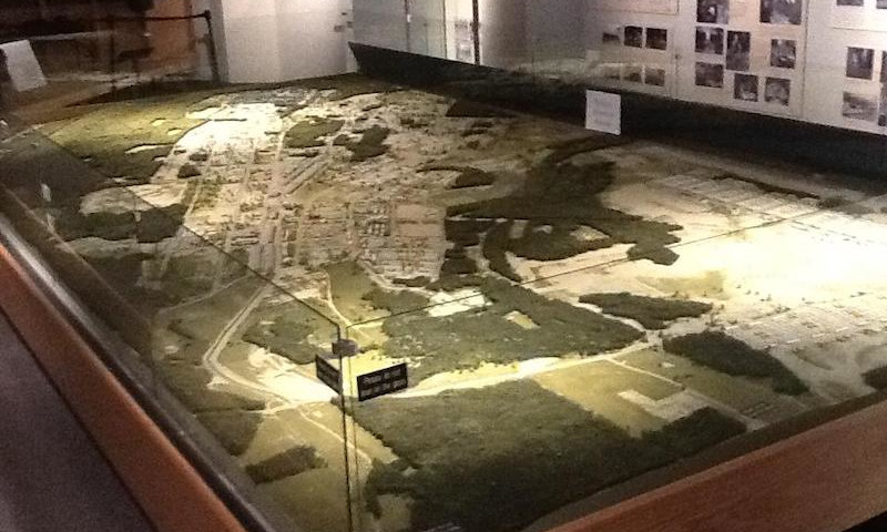 A scale model of a military base is on display and has glass and wood surrounding the model. Historical photos hang on the walls behind the model.
