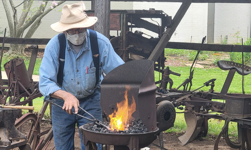 Man in suspenders and hat with facemask navigates tongs while standing at an open-fire pit.