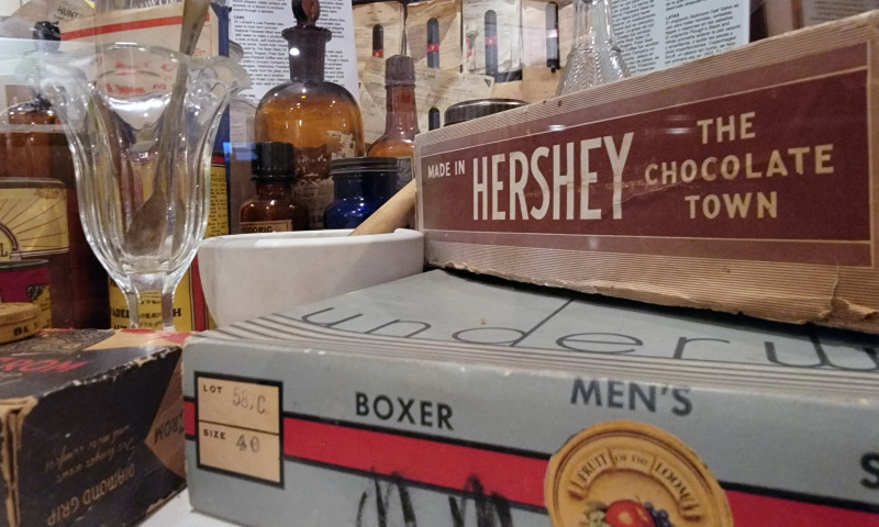 A Hershey's chocolate box sits on top of another box with a bottle and an empty glass with a spoon in the background.