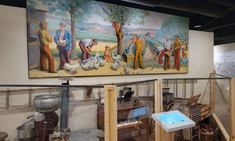 A mural of workers and businessmen standing among crops, trees, and white chickens and roosters over an exhibit of farm implements.