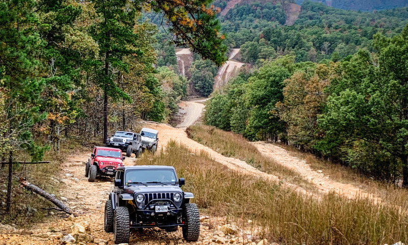 Four off-road vehicles climb a rocky trail, with more trails and mountains in the background.