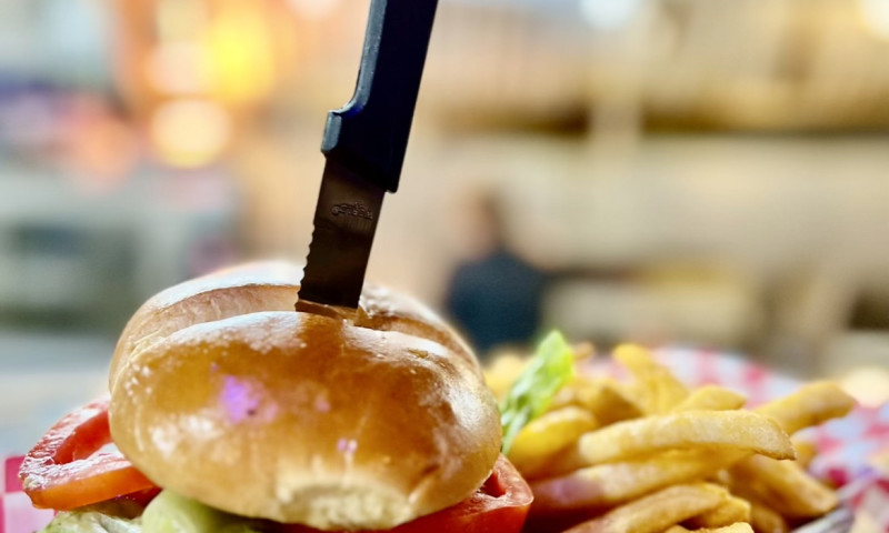 Chicken and Swiss sandwich with knife stuck through the top bun, sits on a metal tray with fries.