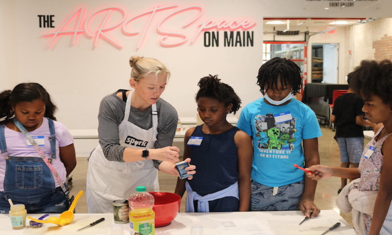 A group of kids stand around a teacher as they learn how to cook. 