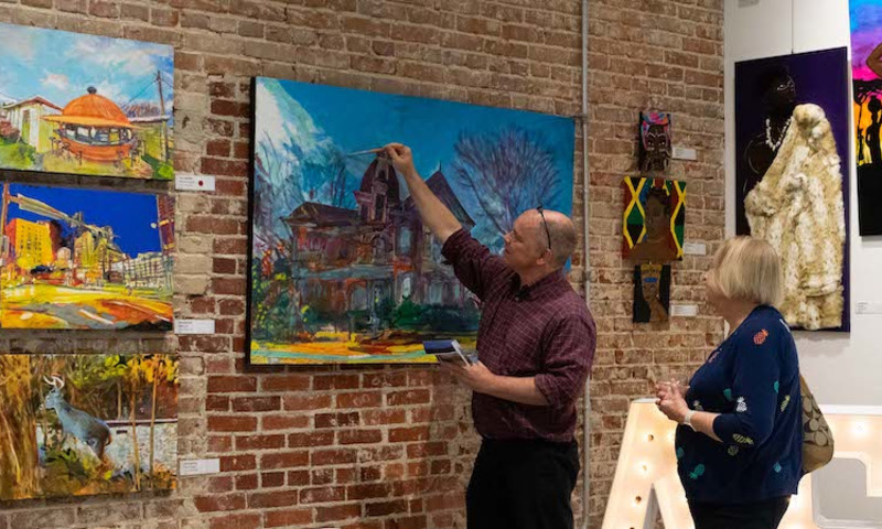 An artist works on touching up his painting that hangs on a brick wall as a woman watches the process.