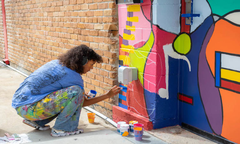 A young woman leans down and paints a mural on a brick wall at ART WORKS on Main in Pine Bluff. 