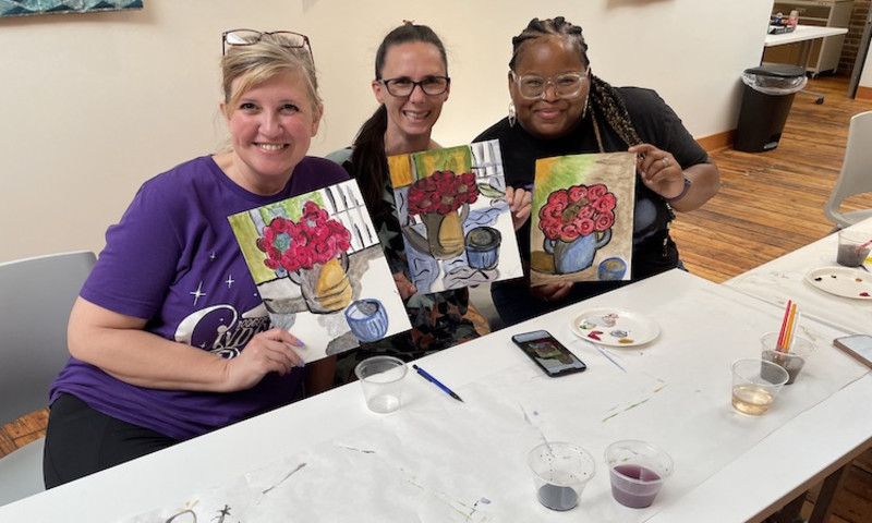 Three women hold up their watercolor artwork they made at the Watercolor & Wine class.