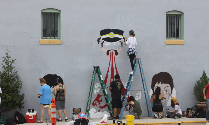 Artists are painting on a wall. In the center, an artist on a ladder is painting a mural reminiscent of Yellow Submarine. To either side, other artists are working on stylized portraits of John and Paul. 