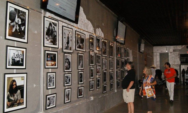 Different photos of the Beatles are framed and hanging on a gallery wall. In the foreground, a color photo of George Harrison is clearly visible. A group of onlookers is admiring the collection. 