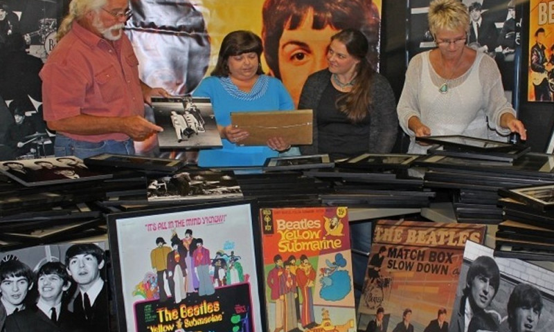 Iconic Beatles album covers populate the foreground, with Yellow Submarine front and center. A group of museum visitors are browsing through stacks of framed Beatles photos. A man is holding a photo and telling a story to others. 