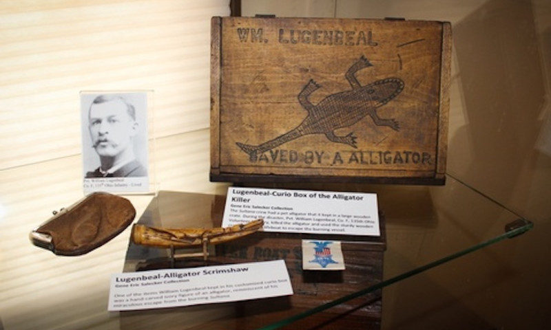 A wooden curio case is on display. In it is carved a crude alligator and the message “saved by a alligator” beneath the name of the original owner, Private William Lugenbeal. A photo of the owner, coin purse and other artifacts are also on display.